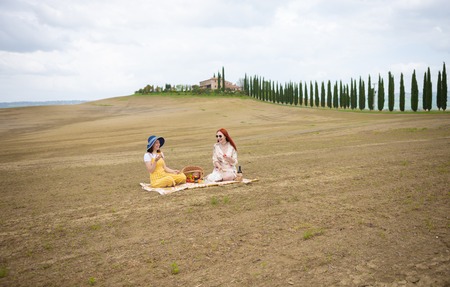 Two women sitting on the blanket having a picnic eating fruits on the background of the cypress treesの写真素材