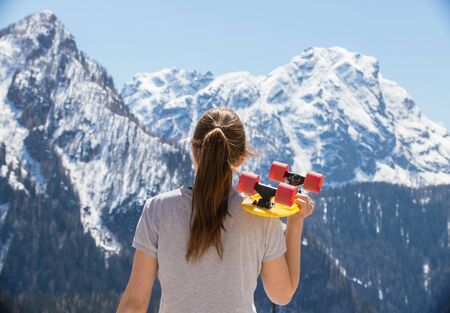 A young woman with blonde hair standing on a background of mountains and holding a skateboardの写真素材