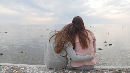 Two young women sitting on the waterfront. Huging and look awayの写真素材