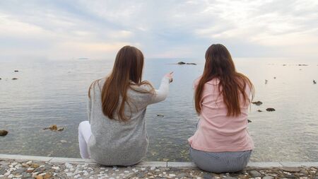 Two young women sitting on the waterfront and show each other birds in seaの写真素材