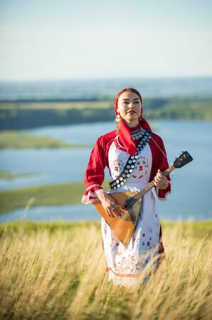 A young woman in russian folk clothes standing on the field holding balalaikaの写真素材