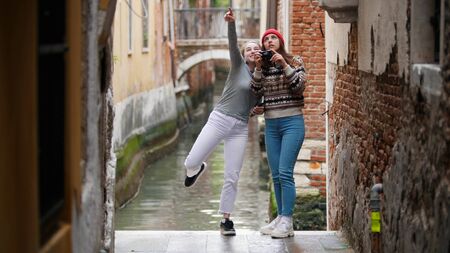 Two young women travelers in warm clothes standing on a background of a water canal and taking photos - Venice, Italyの写真素材