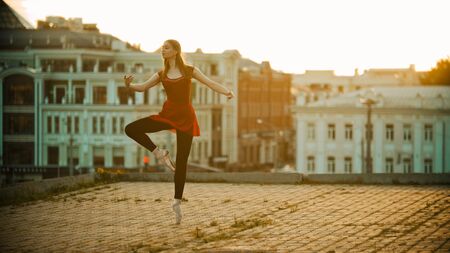 Young inspired woman in red dress ballerina standing in the graceful pose on the roof - modern buildings on the background - bright sunsetの写真素材