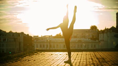 Young woman ballerina training on the roof - standing in the pose showing her stretching - raised her leg up - bright sunsetの写真素材