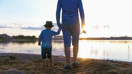 Father and son in striped shirts with fishing rods staying on the riverside at sunsetの写真素材