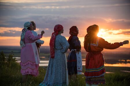 People in traditional russian clothes standing on the field and looking at the bright sunsetの写真素材