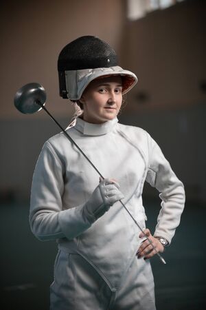 A young woman fencer with an open helmet on standing in the gymの写真素材
