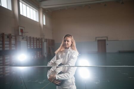 A portrait of a young woman fencer holding her helmet at the school gym