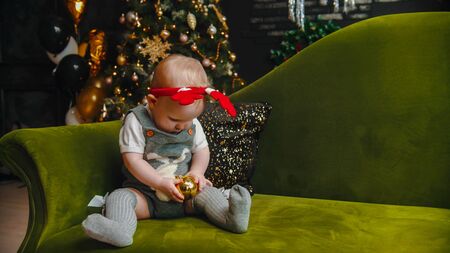 Christmas concept - baby is sitting on sofa and playing with christmas ball