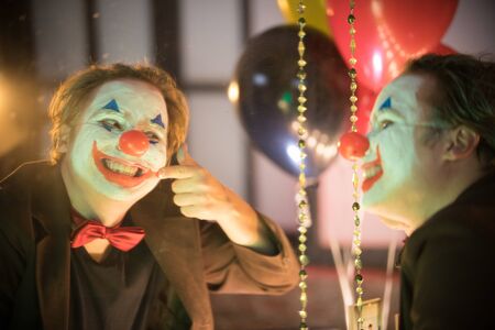 Clown concept - a crazy man clown in the dressing room applying the paint - drawing a smile with a...