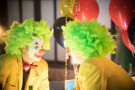 Clown concept - a lost man clown in yellow jacket and green wig looking at himselfの写真素材