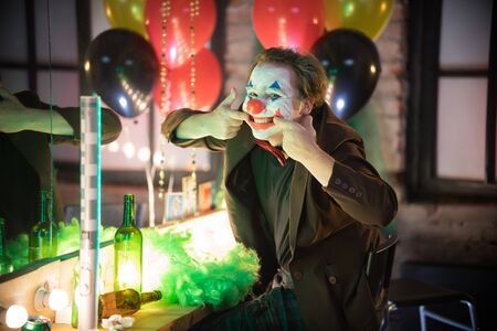 Clown in the dressing room concept - a crazy man clown looking in the camera and stretching his...