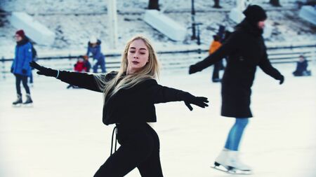 A young blonde attractive woman professional figure skater skating on the outdoors ice rink around peopleの写真素材