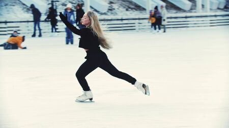 A young blonde attractive woman figure skater skating on the outdoors ice rink around peopleの写真素材