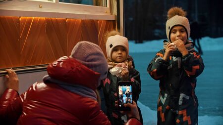A young woman taking photos of her children drinking hot drinks and eating donuts outdoors in winter