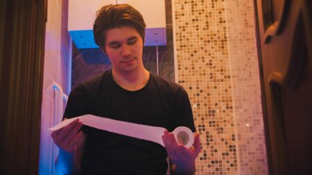 Young man sitting on a toilet and holding toilet paperの写真素材