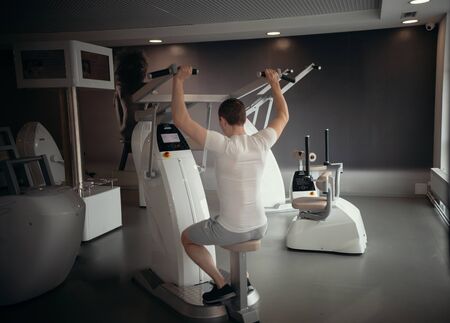 Modern gym - a young man in white t-shirt sitting on the strength training machine and pumping his hands musclesの写真素材