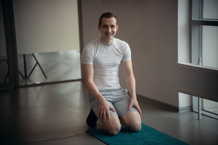 Modern gym - a young man in white t-shirt sitting on the yoga mat