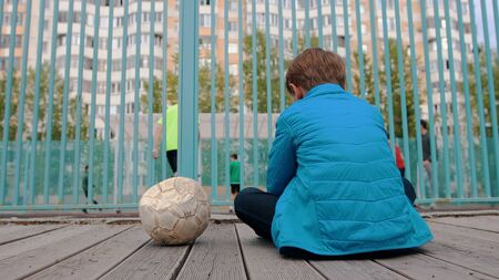 A little boy sitting on the bench near the deflated ball and watching other kids playing footballの写真素材