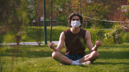 A young man in the medical mask sitting in the park and meditatingの写真素材