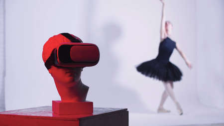 Young woman ballerina in black dress dancing - a head of greek sculpture in VR glasses on the...