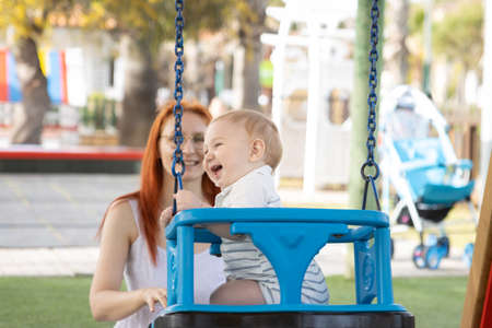 Happy family vacation - funny baby swings on swings in playground and his mom sits near himの写真素材
