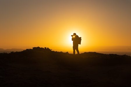 A man stands on top of a cliff and takes photos of the sea on sunsetの写真素材