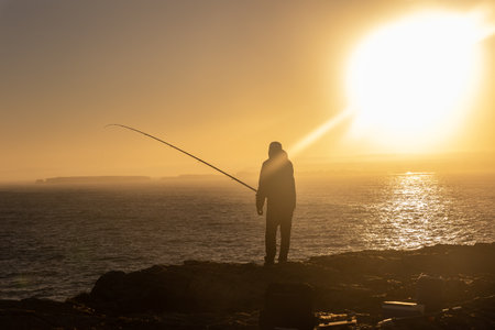 A lonely man standing on the top of the cliff and fishing in the sea at sunsetの写真素材
