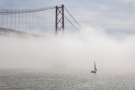 Suspension bridge shrouded in white fog - sailboat sails the sea