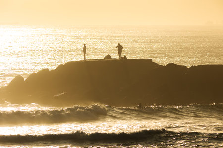 Silhouettes of people standing on a cliff in the middle of the sea and fishing at sunset