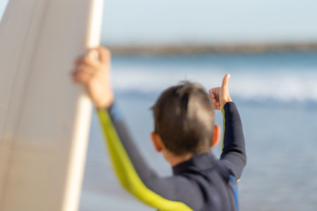 Little boy in wetsuit shows thumbs up looking at surfboard - back viewの写真素材
