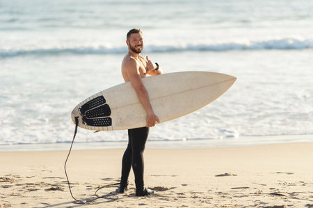 An athletic man surfer with naked torso standing by the sea and showing a thumb up looking in the cameraの写真素材