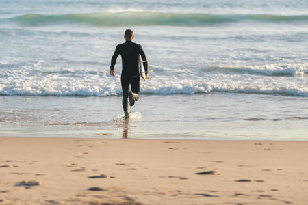 An athletic man surfer in a wetsuit running to the ocean holding a surfing boardの写真素材