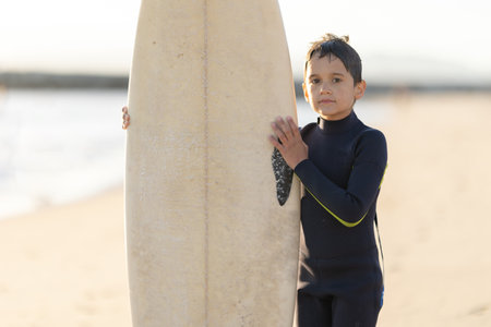 A little boy in a wetsuit holding a surfboard on the seashoreの写真素材