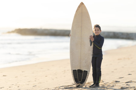 A little boy in a wetsuit holding a big white surfboard standing on the seashoreの写真素材