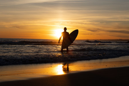 Silhouette of a man holding surfboard standing on the seashore at sunset