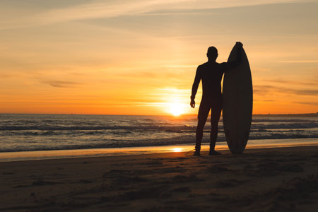 A fit man standing on the seashore leaning on a surfboard at sunsetの写真素材