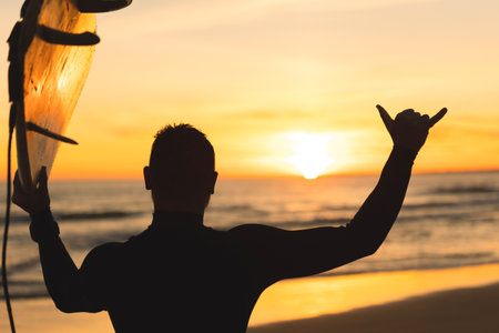 Silhouette of a man holding a surfboard showing shaka at sunsetの写真素材