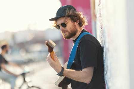Smiling man hipster standing by the wall and playing guitar