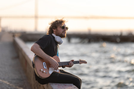 A man hipster sits on the embankment with his legs dangling to the water and playing the guitar