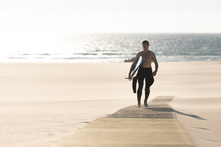 A man with naked torso walking on the path on the beach holding a surfboardの写真素材