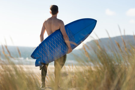 A man standing on the beach holding a blue surfboard - view from the back