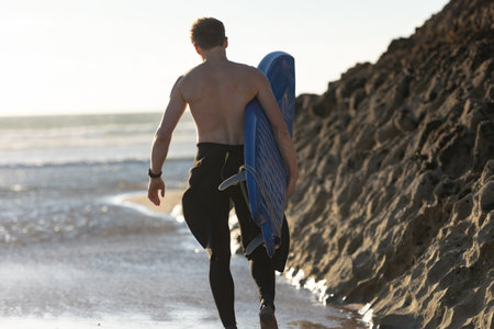 A man with a naked torso standing by a rock holding a surfboard - view from the backの写真素材