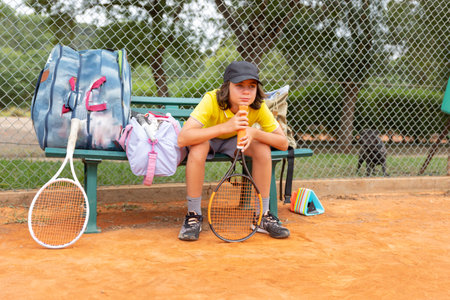 A boy sitting on a bench with sports bafs by the tennis court and waiting for his turnの写真素材