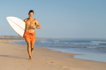 An attractive smiling man with nice athletic body running on the shore holding a surfboard and showing shakaの写真素材