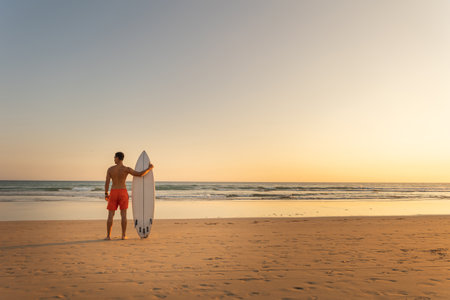 A man with athletic body standing on the shore holding a surfboard at early sunset - view from the backの写真素材