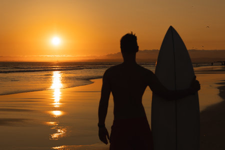 Black silhouette of an athletic man standing on the shore holding a surfboard and looking at sunsetの写真素材
