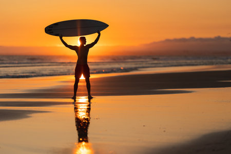Black silhouette of an attractive man on the shore holding a surfboard above his head at orange sunsetの写真素材