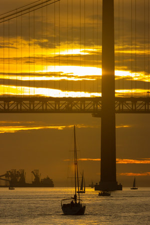 Sailing boat is sailing towards the bridge at bright yellow sunset - oil producing vessel on the...