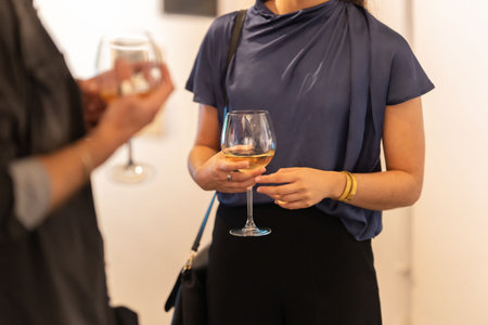 A young woman holding a glass of wine at the art exhibition and talking with her friend